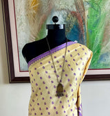 A cream-colored saree with purple borders and small purple motifs, displayed on a black mannequin in an indoor setting with a framed painting and a small plant on a side table.